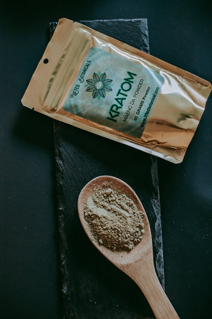 Kratom Maeng Da powder displayed with packet and wooden spoon on a dark surface.